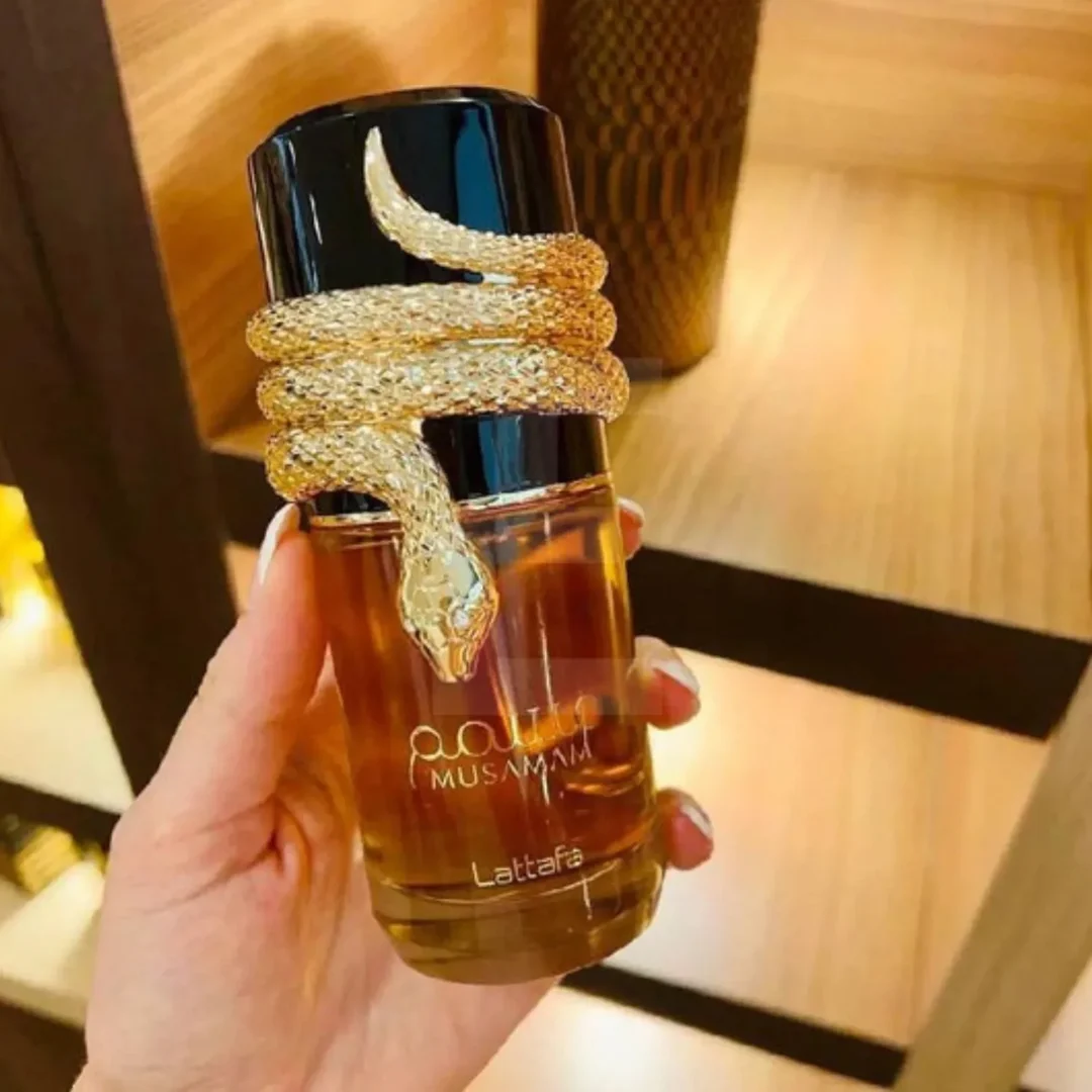Lattafa Musamam Eau De Parfum 100ml by Lattafa — A golden snake coils around a clear glass perfume bottle filled with amber liquid, next to a textured brown cylindrical case with a snake-skin pattern. | Lattafa Musamam Eau De Parfum 100ml by Lattafa — Ular emas melilit botol minyak wangi kaca jernih berisi cecair