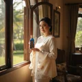 Woman holding Azeezah Lattafa blue glass fragrance near window looking outside smiling