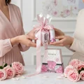 Woman holding Washwasha Lattafa perfume gift set wrapped pink ribbon surrounded roses on table