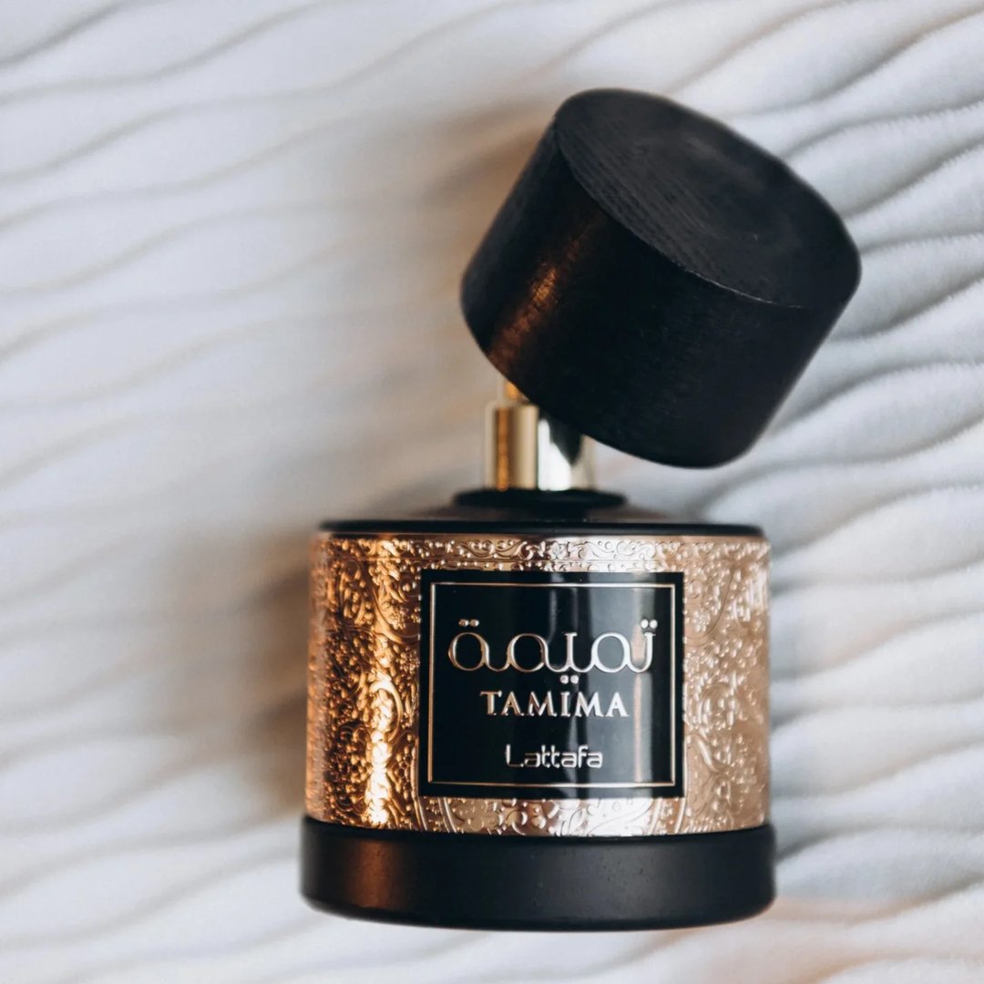 Lattafa Tamima 100ml Eau de Parfum — A black perfume bottle with a wooden cap and a rose gold label sits next to its matching black textured box with rose gold Arabic calligraphy and text. | Lattafa Tamima 100ml Eau de Parfum — Botol minyak wangi hitam dengan penutup kayu dan label emas mawar duduk bersebelahan dengan