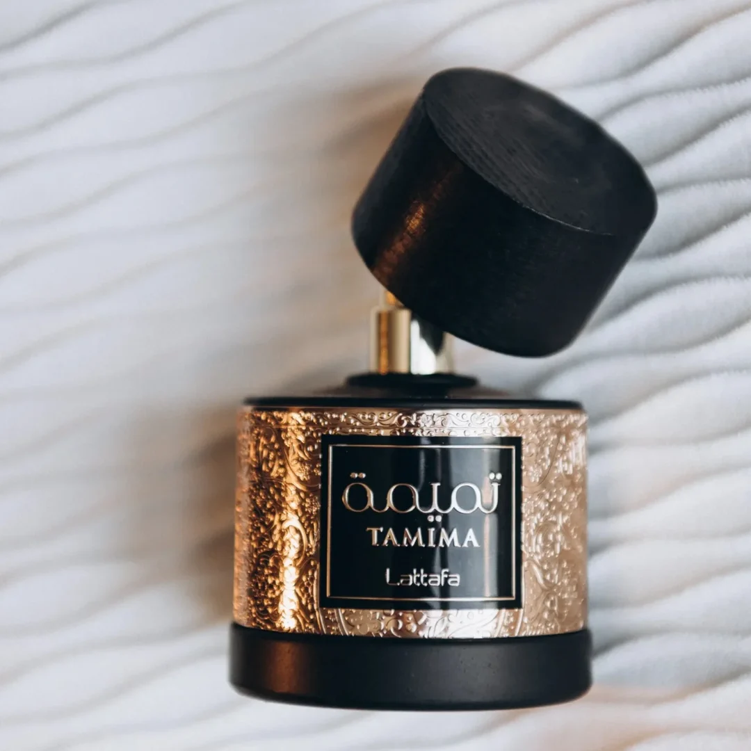 Lattafa Tamima 100ml Eau de Parfum — A black perfume bottle with a wooden cap and a rose gold label sits next to its matching black textured box with rose gold Arabic calligraphy and text. | Lattafa Tamima 100ml Eau de Parfum — Botol minyak wangi hitam dengan penutup kayu dan label emas mawar duduk bersebelahan dengan